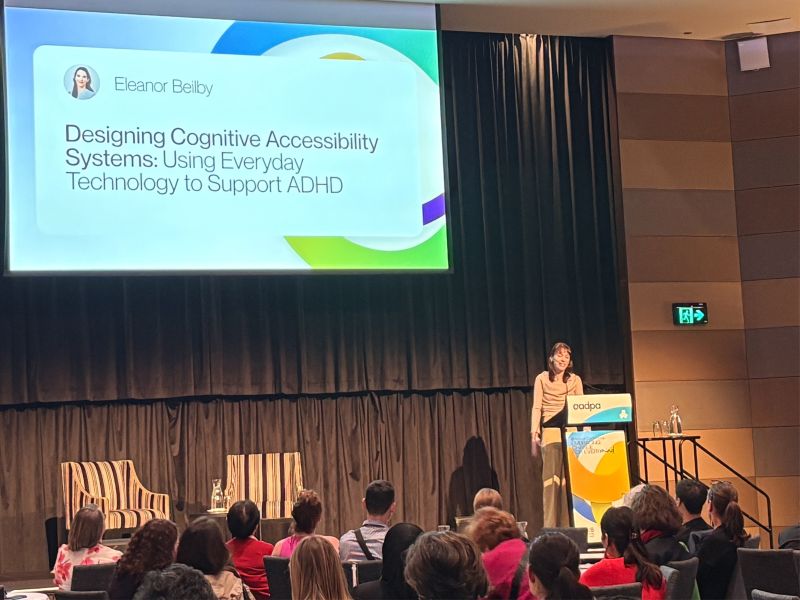 Eleanor Beilby presenting at the lectern at the AADPA 9th Annual Conference, with her talk title slide projected behind her