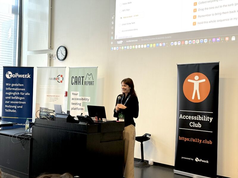 Eleanor Beilby speaking with a microphone beside the Accessibility Club banner at the Accessibility Club Summit 2026 in Würzburg